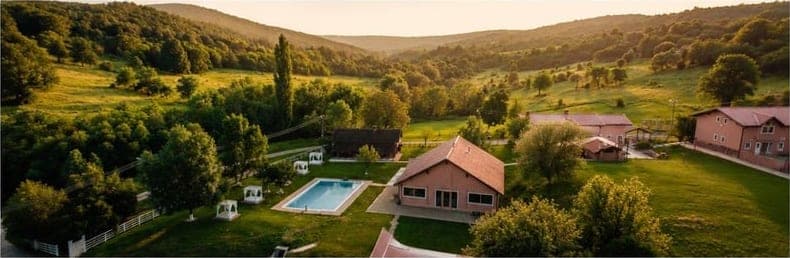 Curtea resortului văzută de sus: piscina cu șezlonguri, casele cu acoperiș roșu și pajiștea din jur.